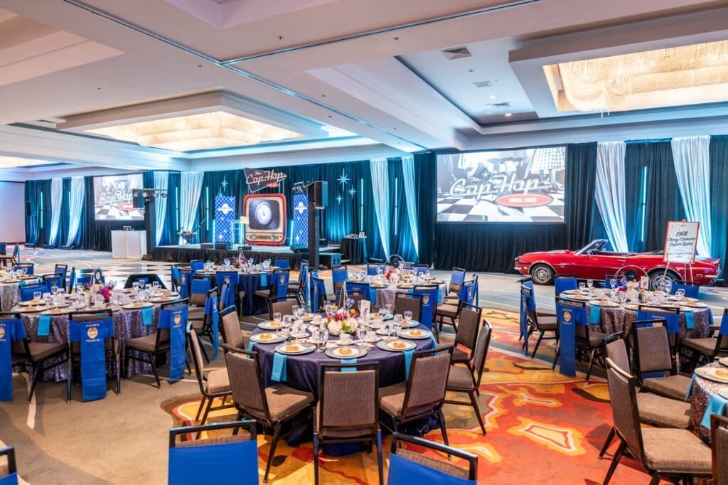 Retro-themed Cop Hop gala ballroom with dinner tables, custom stage design, projection screens, and a classic red convertible.