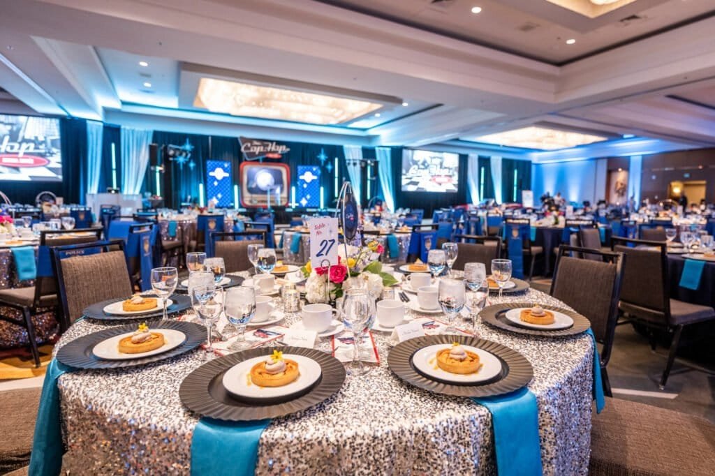 Silver sequin dinner table set with blue accents and florals in front of the retro-themed Cop Hop gala stage.