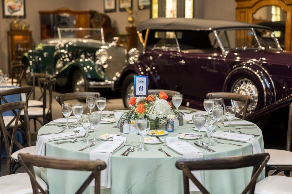 Dinner table with floral centerpiece set among vintage automobiles inside an automotive museum event venue.