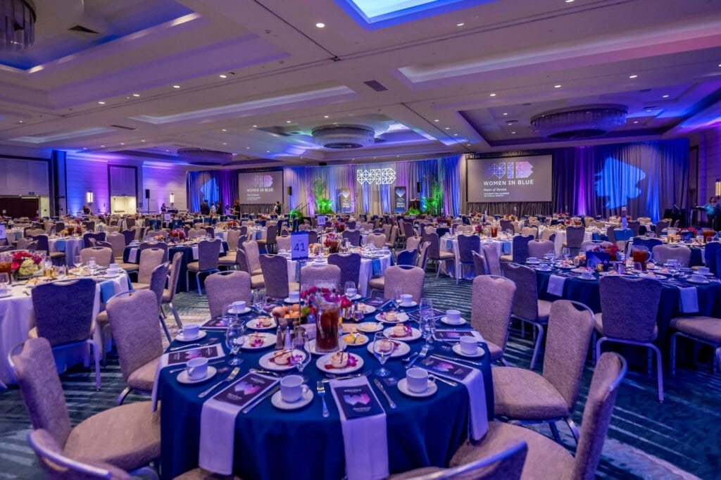 Women in Blue luncheon ballroom with blue table settings, floral centerpieces, stage screens, and purple-blue event lighting.